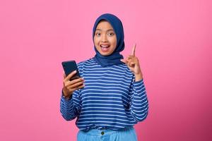 ritratto di una giovane donna asiatica eccitata che tiene in mano uno smartphone con un'idea o una domanda che punta il dito foto