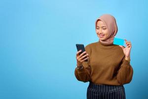 felice giovane donna asiatica che mostra la carta di credito e il telefono cellulare a portata di mano su sfondo blu foto