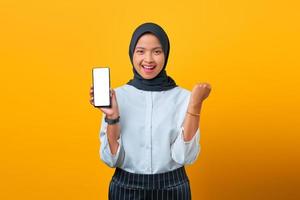 donna asiatica felice che mostra lo schermo vuoto del telefono cellulare e il successo del gesto della mano su sfondo giallo foto