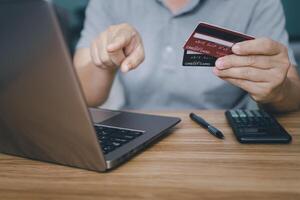 un' uomo hold credito carta e punto per tenere sotto controllo il computer portatile computer nel mano accedere carta numero con calcolatrice su legna tavolo per calcolare i soldi di il mese foto