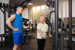 giovane bello allenatore uomo aiuta un' pensionato per fare un esercizio su il simulatore nel il palestra. contento persone Sorridi. attivo salutare gli sport stile di vita, anziano concetto. tavoletta foto