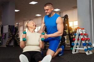 giovane maschio allenatore aiuta un vecchio donna esercizio nel il palestra. attivo, sportivo, salutare stile di vita. anziano concetto. interno. contento sorridente le persone, la pensione età. manubri, muscolo swing foto