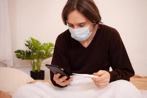 un' malato uomo chiamate un ambulanza su il Telefono. un' mascherato uomo controlli il temperatura e quadranti il medico numero su il Telefono. coronavirus, casa quarantena. foto