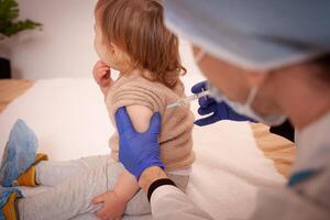 il medico vaccina il bambino contro coronavirus. il bambino è pianto e paura. un' uomo nel un' veste, cappello, maschera e guanti fa un iniezione per il bambino. casa quarantena foto