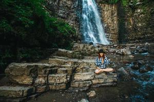 donna con i capelli rossi in un cappello e una camicia meditando sulle rocce nella posizione del loto contro una cascata foto