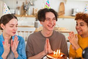 rendere un' desiderio. uomo indossare festa berretto soffiaggio su ardente candele su compleanno torta. contento compleanno festa. gruppo di amici auguri tipo contento compleanno. persone festeggiare compleanno con festa a casa foto