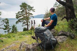 escursioni a piedi arrampicata scogliera avventura. zaino in spalla uomo riposo dopo escursioni a piedi guardare a bellissimo Visualizza. escursionista con zaino si siede su grande roccia sopra verde foresta e lago. indietro Visualizza escursionista uomo godere escursione turismo foto