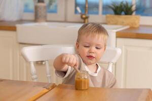 contento famiglia a casa. bambino ragazzo alimentazione lui stesso nel cucina. poco ragazzo con disordinato divertente viso mangia salutare cibo. bambino impara mangiare di lui stesso Tenere cucchiaio. se stesso alimentazione foto