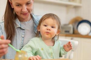 contento famiglia a casa. madre alimentazione sua bambino ragazza a partire dal cucchiaio nel cucina. poco bambino piccolo bambino con disordinato divertente viso mangia salutare cibo a casa. giovane donna mamma dando cibo per ragazzo figlia foto