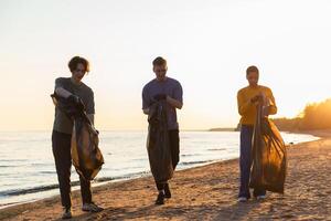 terra giorno. volontari attivisti squadra raccoglie spazzatura pulizia di spiaggia costiero zona. donna mans con spazzatura nel spazzatura Borsa su oceano costa. ambientale conservazione costiero zona pulizia foto