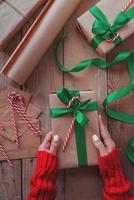 mani di donna che tengono un regalo di Natale avvolto con carta artigianale, nastro verde e bastoncino di zucchero vista dall'alto sul tavolo di legno foto