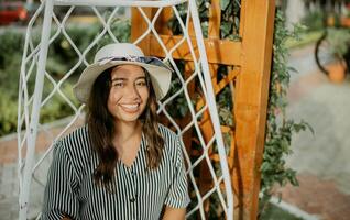 ritratto di un' sorridente ragazza nel un' cappello seduta su un' bianca swing nel un' giardino. stile di vita di ragazza nel cappello seduta su un' swing guardare a telecamera foto