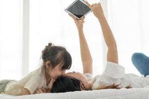 ragazzo asiatico che bacia sua madre mentre la madre usa il tablet per scattare loro foto sul letto. concetto di famiglia insieme