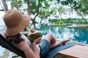 giovane donna che indossa cappello di paglia e abbigliamento casual seduto sulla panchina vicino alla piscina e bere succo di cocco durante le vacanze estive