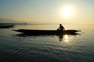 sul lato del lago, pescatore asiatico seduto sulla barca e usando la canna da pesca per catturare i pesci all'alba foto