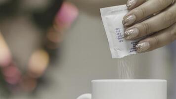 vicino su di un' donna mani lancio zucchero in un' bianca boccale con caldo bevanda su sfocato sfondo. azione filmato. femmina mani aggiungendo bianca zucchero in tè o caffè. foto