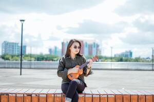 giocando ukulele di giovane bellissimo asiatico donna indossare giacca e nero jeans in posa all'aperto foto