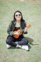 giocando ukulele di giovane bellissimo asiatico donna indossare giacca e nero jeans in posa all'aperto foto