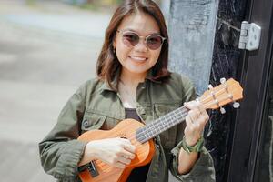 giocando ukulele di giovane bellissimo asiatico donna indossare giacca e nero jeans in posa all'aperto foto