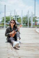 giocando ukulele di giovane bellissimo asiatico donna indossare giacca e nero jeans in posa all'aperto foto