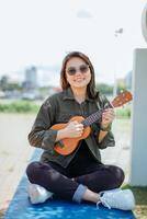 giocando ukulele di giovane bellissimo asiatico donna indossare giacca e nero jeans in posa all'aperto foto