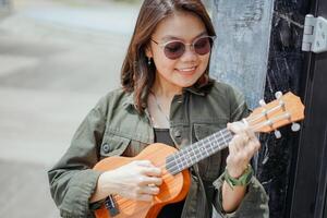 giocando ukulele di giovane bellissimo asiatico donna indossare giacca e nero jeans in posa all'aperto foto