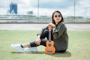 giocando ukulele di giovane bellissimo asiatico donna indossare giacca e nero jeans in posa all'aperto foto