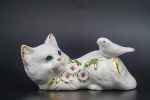 porcellana giocoso gatto, ceramica statuetta. colomba, abbellimento, petali e le foglie dorato bordi. dipinto a mano raffinato. brillante superficie, da collezione elemento. grazioso postura, fatto a mano. nero sfondo foto