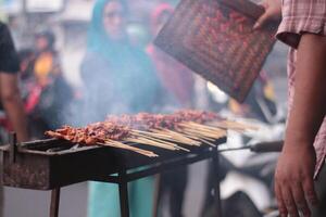 pollo satay al di sopra di tradizionale carbone fuoco. il satay ustioni con Fumo e un appetitoso aspetto esteriore. mani cucinando satay su il griglia. persone siamo già fare la fila. foto