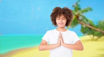 giovane donna afro meditando sulla spiaggia con gli occhi chiusi. asana yoga con gli occhi chiusi. donna durante la meditazione con le mani namaste meditazione nella natura mare foto