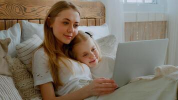 piccolo poco figlia con caucasico mamma donna madre rilassare nel letto insieme affettuoso dire bugie nel Camera da letto con il computer portatile video chiamata conferenza parlare Guardando film film in linea cartone animato nel Internet parlando foto