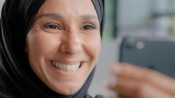 colpo alla testa giovane bella contento arabo donna nel hijab sorridente con bianca salutare denti guardare a smartphone schermo gioia a bene notizia Guardando divertente video su telefono riceve positivo risultato di e-mail foto