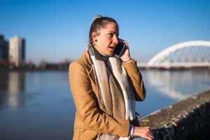 sorpreso donna parlando su un' mobile Telefono nel caldo capi di abbigliamento e riposo di il fiume su un' soleggiato inverno giorno. foto