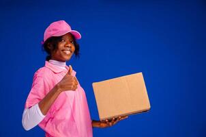 bellissimo africano femmina Corriere lavoratore Tenere un' scatola, fa un' pollici su gesto foto