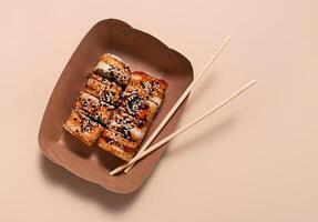 asiatico Sushi. Sushi con anguilla, salsa e sesamo nel un' carta contenitore su un' leggero sfondo. cibo consegna. lato Visualizza. copia spazio. foto
