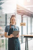 bellissimo giovane barista donna nel grembiule Tenere tavoletta e in piedi nel davanti di il porta di bar con Aperto cartello tavola. attività commerciale proprietario avviare, pmi imprenditore venditore attività commerciale concetto. foto