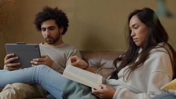 giovane donna di razza mista e giovane uomo mediorientale seduto sul divano, la donna legge il libro, l'uomo tiene il tablet, parlando seriamente foto