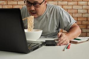 lavoratore di sesso maschile tailandese impegnato a lavorare con il laptop, usa le bacchette per mangiare frettolosamente noodles istantanei durante la pausa pranzo in ufficio, perché veloce, gustoso ed economico. nel tempo fast food asiatico, stile di vita malsano. foto