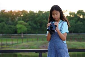 asiatico ragazza usi binocolo per osservare il natura, uccelli, e animali di il nazionale parco mentre campeggio con sua famiglia. con il concetto di apprendimento al di fuori il aula natura studia, esplorazione. foto