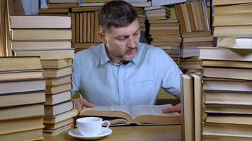 un' barbuto uomo legge un' libro nel il biblioteca. pile di libri e tazza di tè su tavolo. concetto di lettura e formazione scolastica. foto