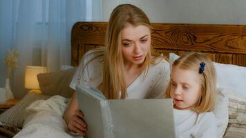 famiglia apprendimento concetto. caucasico madre Aiuto poco piccolo carino figlia prescolastico bambino ragazza bambino lettura libro nel letto a casa Camera da letto mamma con ragazzo leggere letteratura Fata racconto elementare formazione scolastica foto