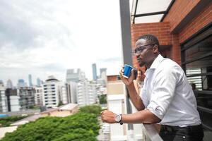 africano attività commerciale inteligente uomo sta a all'aperto terrazza costruzione. persone con su mano nel bene sensazione con città spazio costruzione. rilassante di persone attività commerciale mattina. foto