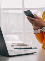 attività commerciale donna utilizzando mobile Telefono durante Lavorando su il computer portatile computer, fare surf il Internet, ricerca attività commerciale dati a moderno ufficio. asiatico donna d'affari in linea Lavorando su computer a posto di lavoro foto