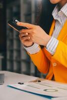 attività commerciale donna utilizzando mobile Telefono durante Lavorando su il computer portatile computer, fare surf il Internet, ricerca attività commerciale dati a moderno ufficio. asiatico donna d'affari in linea Lavorando su computer a posto di lavoro foto