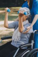 giovane infermiera o fisioterapista nel scrub porzione un' contento pensionato vecchio donna fare fitness esercizi con leggero peso manubri a casa. concetto di fisioterapia per gli anziani foto