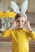 bellissimo poco ragazzo indossare un' coniglietto costume, giocando nel il cucina, Tenere bianca chiken uova, festeggiare Pasqua vacanza foto