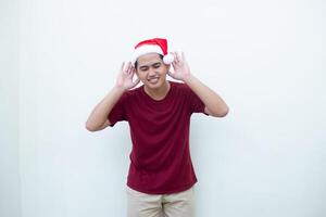 giovane asiatico uomo indossare un' Santa Claus cappello esprimendo ascoltando con mano per orecchio isolato di un' bianca sfondo per visivo comunicazione foto