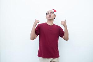 giovane asiatico uomo indossare un' Santa Claus cappello sorridente, shock e puntamento per sua lato isolato di bianca sfondo per visivo comunicazione foto