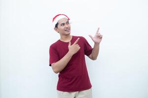 giovane asiatico uomo indossare un' Santa Claus cappello sorridente, shock e puntamento per sua lato isolato di bianca sfondo per visivo comunicazione foto