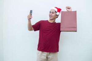 giovane asiatico uomo indossare un' Santa Claus cappello Tenere un' smartphone e un' shopping Borsa con espressioni di Sorridi, shock, e sorpresa, isolato contro un' bianca sfondo per visivo comunicazione foto